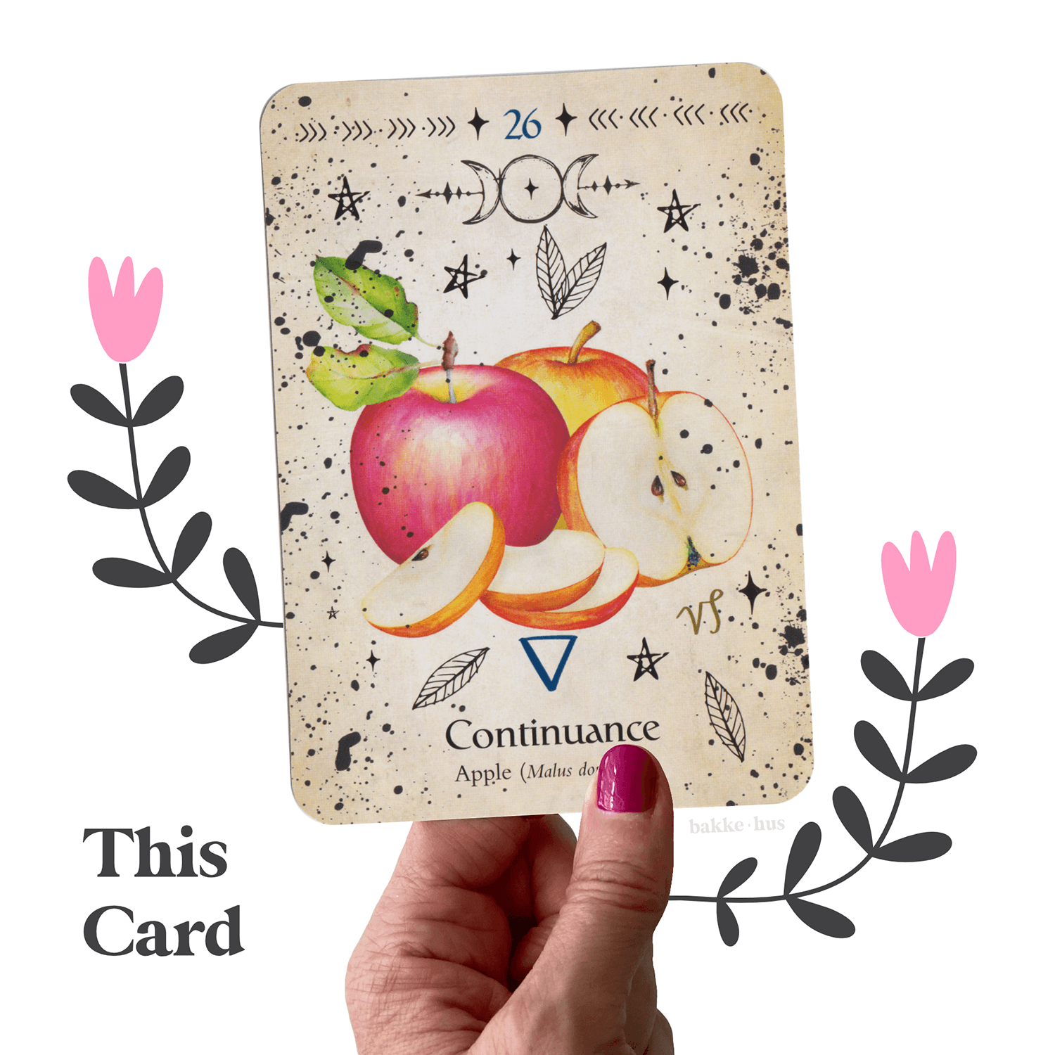 A female hand holding the Apple Oracle Cards from Green Witch Oracle Deck by Cheralyn Darcey. The image also contains two pink & black illustrated flowers and the words 'This Card" and bakkehus logo.