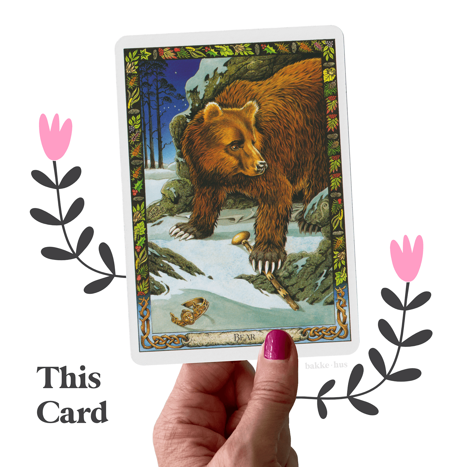 A female hand holding the Bear Oracle cards from Druid Animal Oracle deck by Philip Carr-Gomm The image also contains two pink & black illustrated flowers and the words 'This Card" and bakkehus logo.