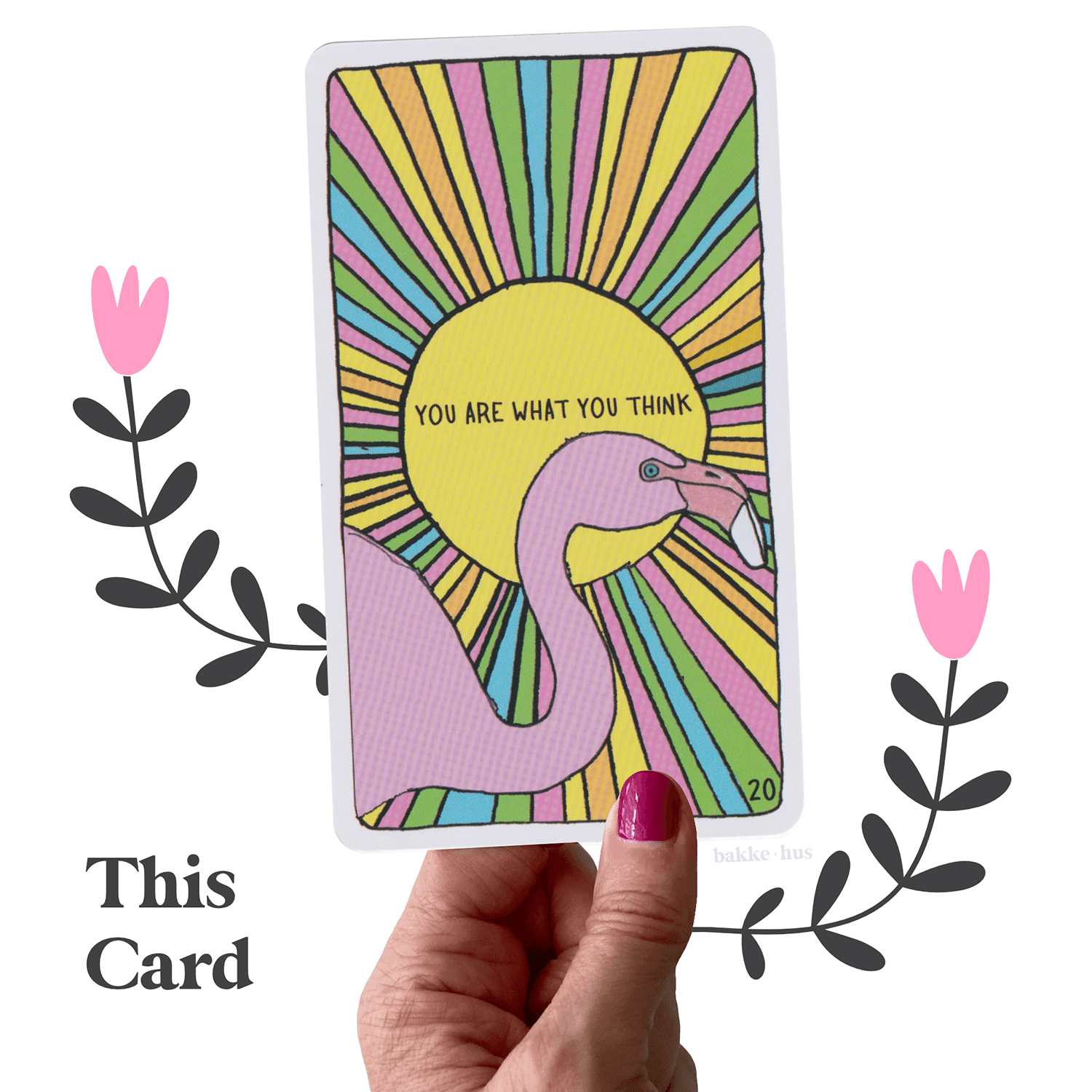 A female hand holding the Flamingo Oracle Card from Animal Apothecary Deck by Cara Elizabeth. The oracle card has the words 'You are what you think' written on it. The image also contains two pink & black illustrated flowers and the words 'This Card" and bakkehus logo.